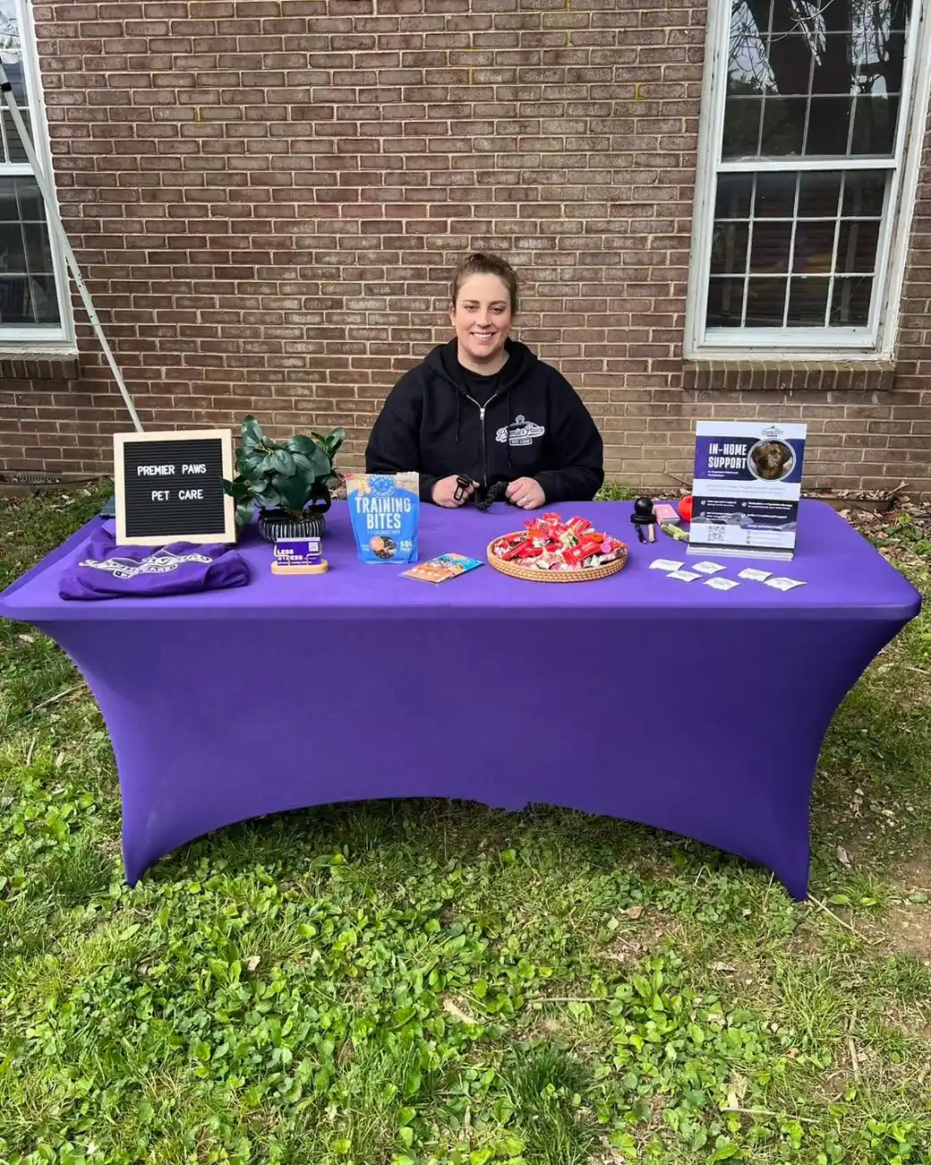 Team member seated at a purple-draped Premier Paws event table with a letterboard, treat basket, and brochures outside a brick building