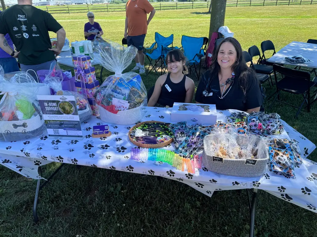 Two team members at an outdoor event table stacked with raffle gift baskets, cookies, and rainbow leashes