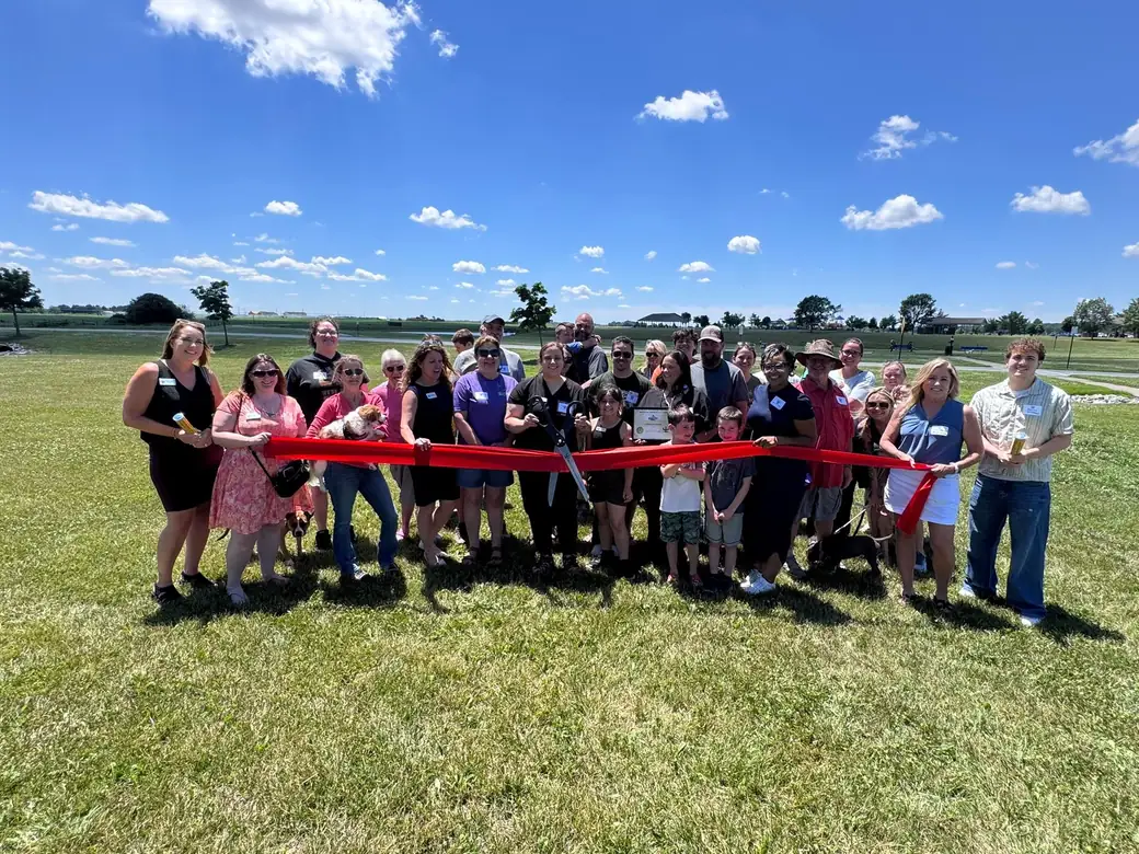 Crowd gathered on a sunny green field for the Premier Paws ribbon-cutting, a red ribbon stretched across the group